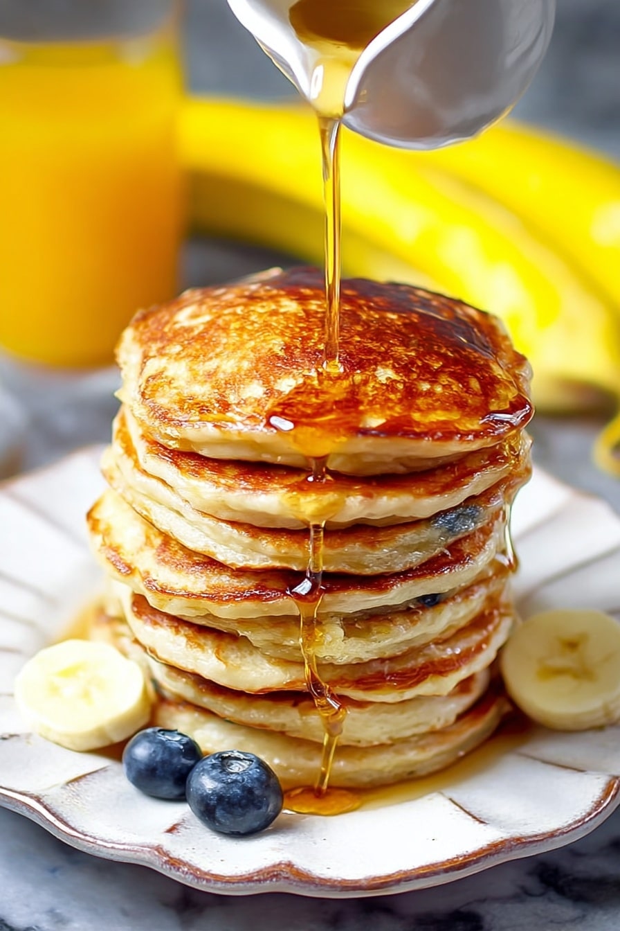 A tall stack of seven thick pancakes with a golden brown color is placed on a white plate with a wavy edge. A golden syrup is being poured from a white small pitcher onto the top pancake, flowing down the sides of the stack. Two fresh blueberries sit beside the pancakes on the plate. In the background, blurred bananas and a glass of orange juice are visible on a white marbled surface. The syrup shine and the pancakes’ cooked texture are clearly visible. Photo taken with an iphone --ar 2:3 --v 7 - Greek Yogurt Pancakes with Lemon, lemon yogurt pancake recipe, fluffy Greek yogurt pancakes, lemon breakfast pancakes, healthy Greek yogurt pancake