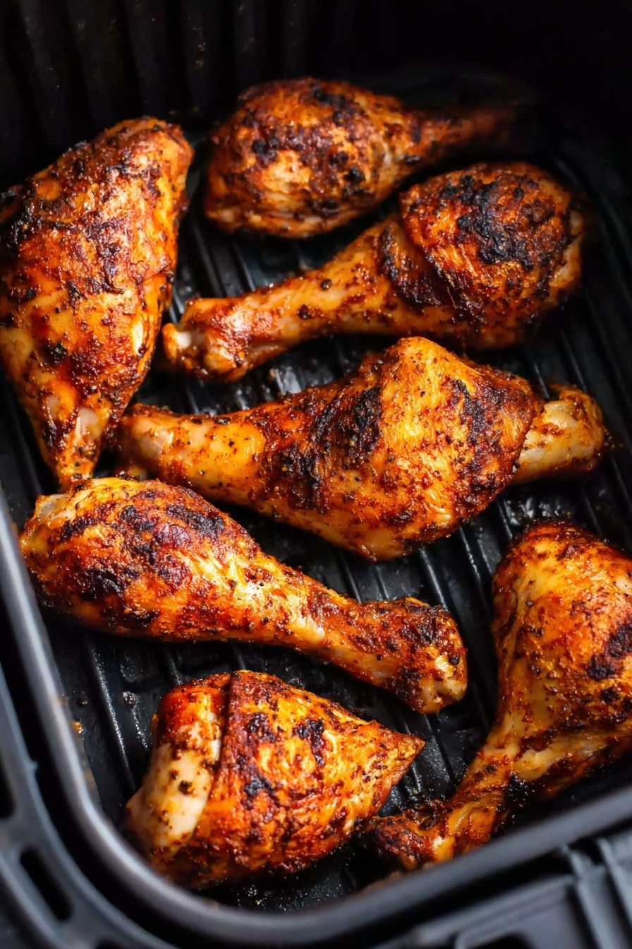 The image shows six browned and crispy chicken drumsticks inside a black air fryer basket. Each drumstick is coated with a dark, spicy seasoning creating a speckled pattern of black, orange, and red spices on their skin. The drumsticks are arranged closely, some slightly overlapping, resting on the ridged black surface of the basket. The texture appears crunchy and well-cooked with a shiny, slightly oily finish. photo taken with an iphone --ar 2:3 --v 7 - Air Fryer Chicken Legs, crispy chicken thighs, quick chicken dinners, healthy air fryer recipes, easy chicken leg recipes