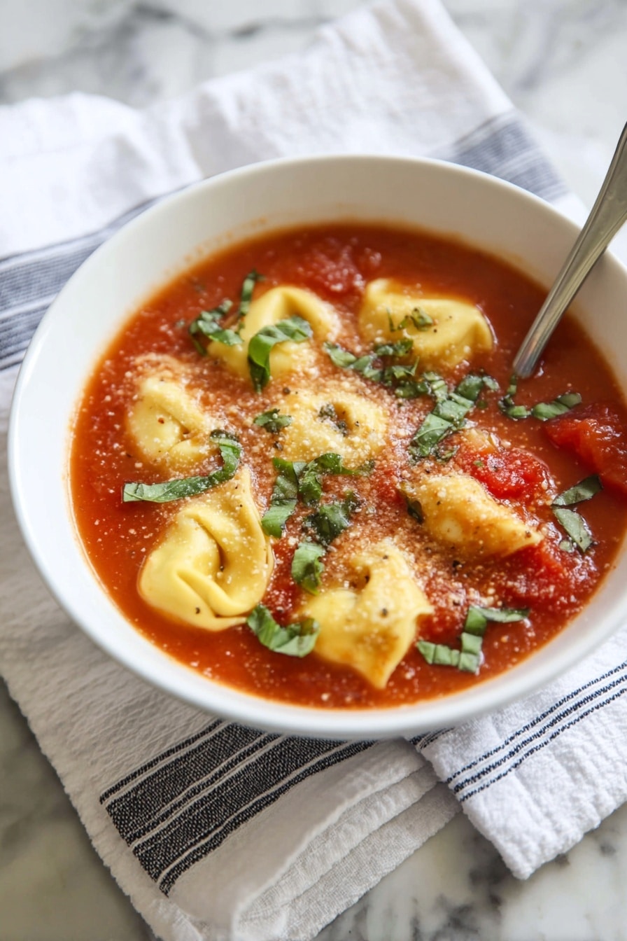 Tomato Basil Tortellini Soup Recipe 8 A white bowl holds a tomato soup base with visible chunks of cooked tomatoes, filled with six yellow tortellini pieces that have a soft, smooth texture lightly bubbling in the warm soup. Fresh green basil strips are scattered on top, adding a fresh contrast to the red soup. A light dusting of grated parmesan cheese covers some of the tortellini and soup surface. A silver spoon partially rests inside the bowl at the back right edge. The bowl sits on a folded white cloth with blue and black stripes, against a white marbled textured surface photo taken with an iphone --ar 2:3 --v 7 - Tomato Basil Tortellini Soup, easy tortellini soup, quick tomato basil soup, comforting pasta soup, hearty vegetarian soup