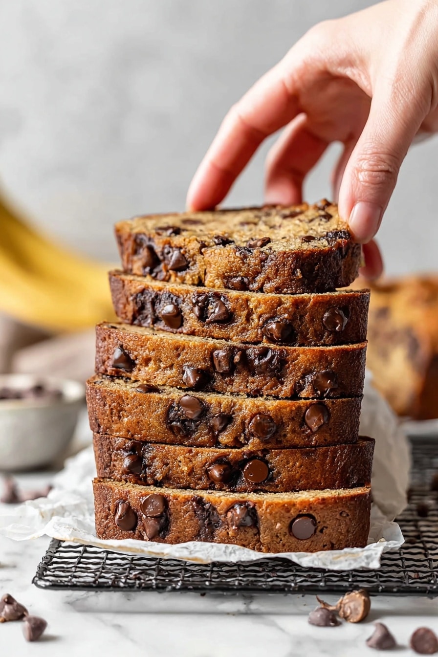 A stack of four thick slices of chocolate chip banana bread resting on white parchment paper on a black wire rack over a white marbled surface. Each slice is dense and golden brown, filled with many dark brown chocolate chips throughout, with some melted and slightly shiny. A woman's hand is lifting the top slice, showing its texture and soft crumb. In the background, there is a blurred yellowish ripe banana on the left side and some more chocolate chip bread slices scattered around, along with a white bowl and some loose chocolate chips in the foreground. Photo taken with an iphone --ar 2:3 --v 7 - Moist Chocolate Chip Banana Bread, banana bread with chocolate chips, easy banana bread recipe, moist banana bread, best chocolate chip banana bread