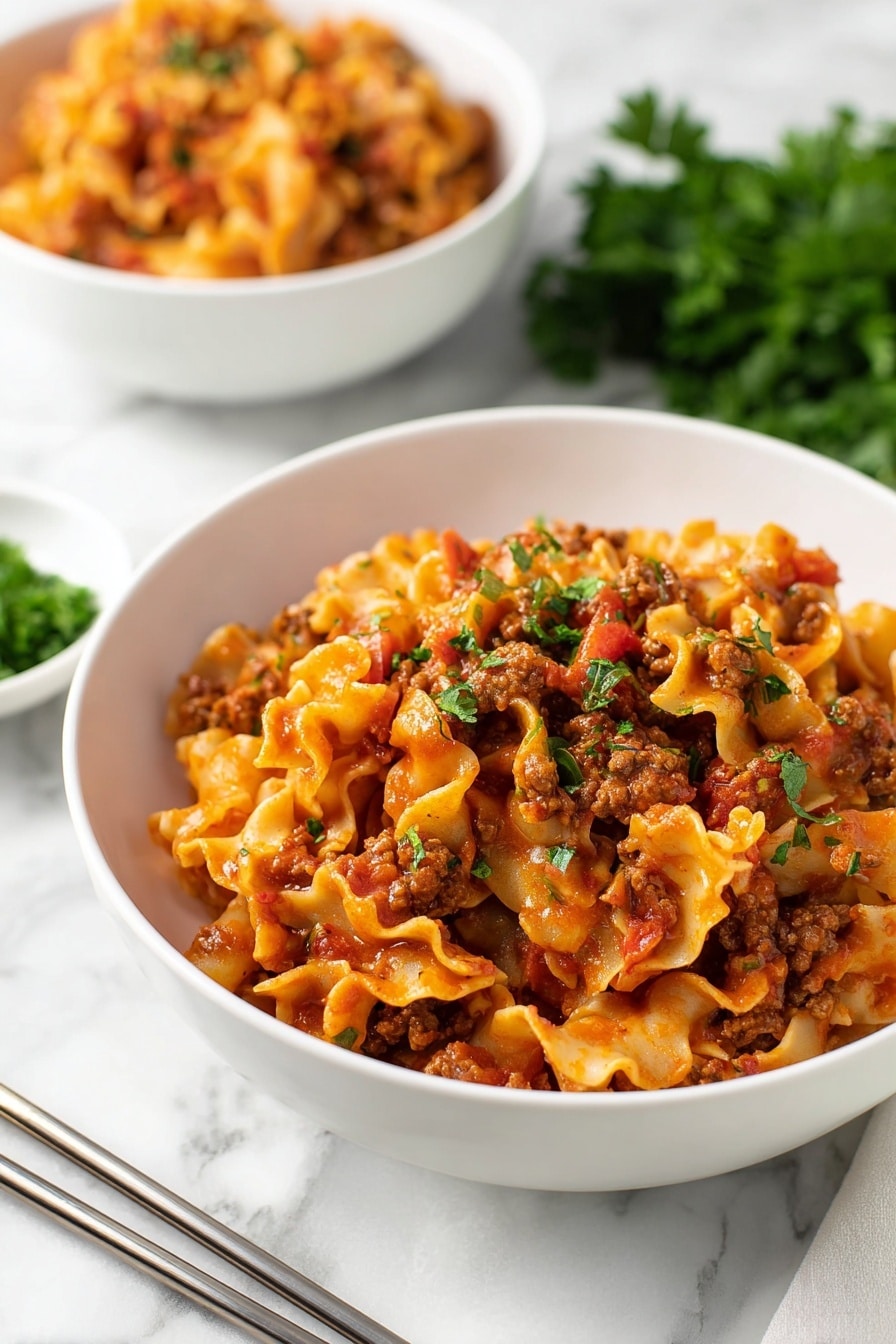 Instant Pot Lazy Lasagna Recipe 8 The image shows a white bowl filled with a layered pasta dish, featuring curly, ruffled-edged noodles mixed with a chunky red tomato sauce and browned ground meat. The pasta is coated in the sauce, giving it a rich orange-red color, with bits of tomato and meat spread evenly throughout. Small green parsley pieces are sprinkled on top for color contrast. Behind the main bowl, there is a second bowl with a similar pasta dish slightly out of focus. The setting includes metal chopsticks resting on a white marbled surface, with some fresh green parsley in the foreground and a blurred bowl of chopped herbs in the background. photo taken with an iphone --ar 2:3 --v 7 - Instant Pot Lazy Lasagna, quick lasagna dinner, easy lasagna recipe, pressure cooker lasagna, weeknight lasagna