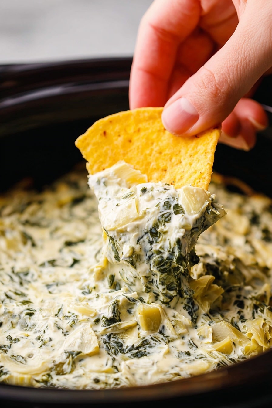 A close-up view of a yellow corn chip being dipped into a thick, creamy dip full of chopped spinach and artichoke pieces. The dip is pale off-white with green spinach bits and small chunks of pale yellow artichoke, filling the inside of a dark bowl with a smooth edge. A woman's hand is holding the chip from above, with fingers visible and a soft light highlighting the textures. The background is blurred but changes to white marbled texture as per instructions. photo taken with an iphone --ar 2:3 --v 7 - Creamy Spinach Artichoke Dip, spinach artichoke dip recipe, crockpot spinach artichoke dip, easy appetizer dip, cheesy spinach artichoke dip