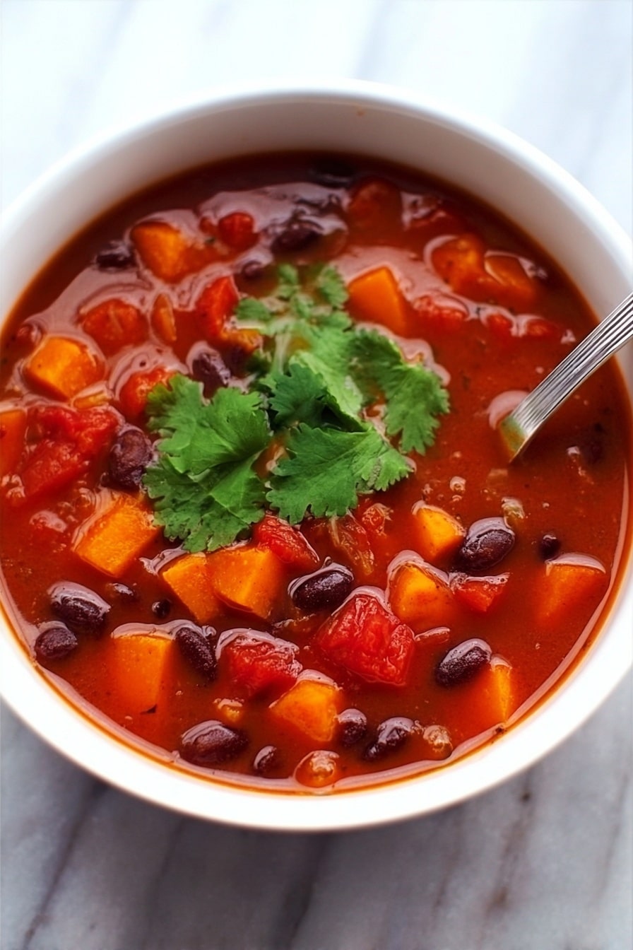 A white bowl filled with thick red soup containing many small pieces. The soup has bright orange cubes, dark beans, and red tomato chunks. On top, there are fresh green cilantro leaves in the center. A shiny metal spoon is placed inside the bowl, resting on the right edge. The background is a white marbled surface. photo taken with an iphone --ar 2:3 --v 7 - Black Bean and Sweet Potato Soup, hearty vegetarian soup, healthy black bean soup, easy sweet potato soup, comforting plant-based soup