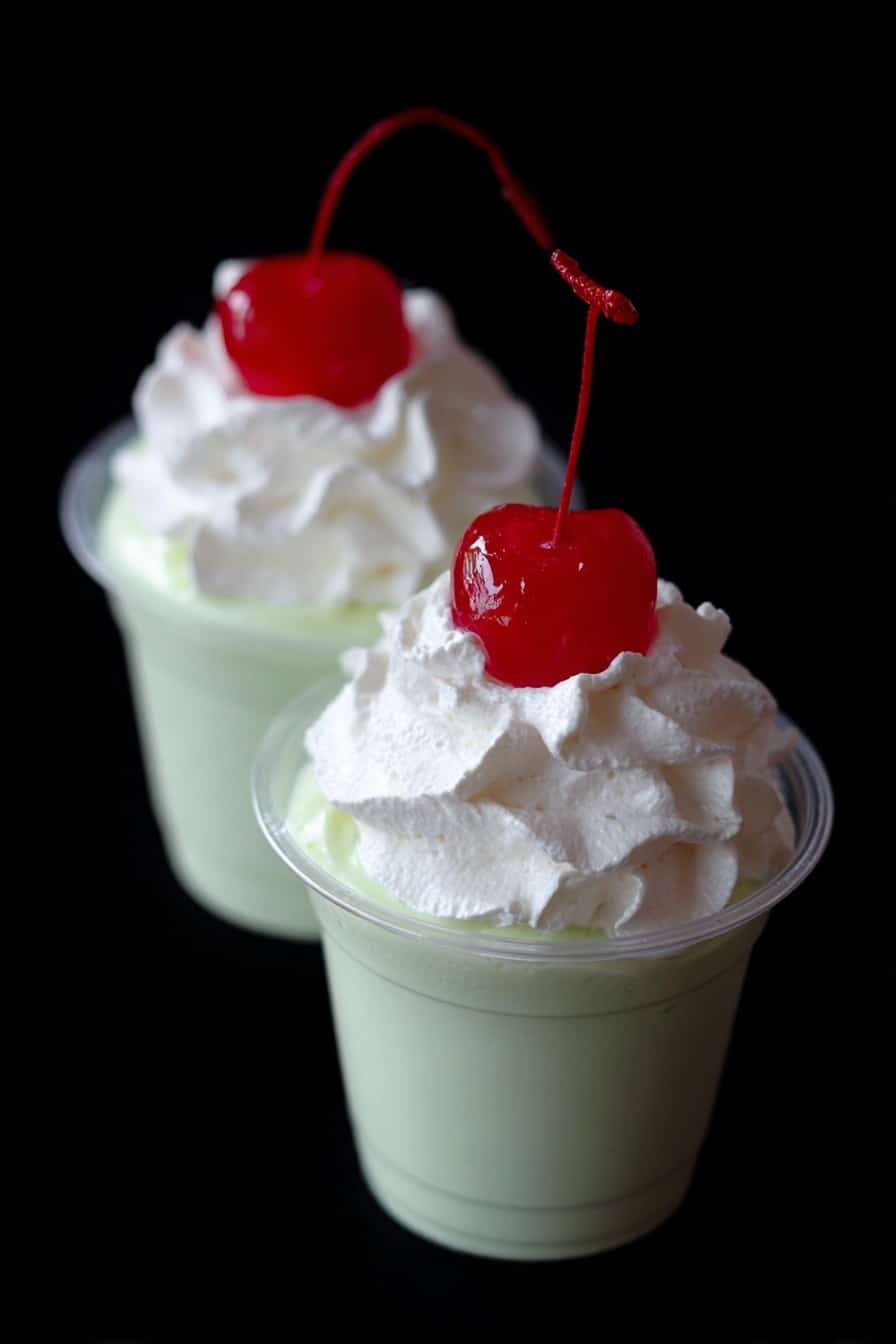 Shamrock Mint Pudding Shots Recipe 8 Two small clear cups filled with a smooth light green creamy layer, topped with a thick dollop of white whipped cream. Each cup has a bright red cherry with a long stem placed on top of the whipped cream. The background is black, making the colors of the dessert stand out clearly. The photo taken with an iphone --ar 2:3 --v 7 - Shamrock Mint Pudding Shots, mint pudding shots, St. Patrick's Day dessert, boozy pudding shots, easy party desserts