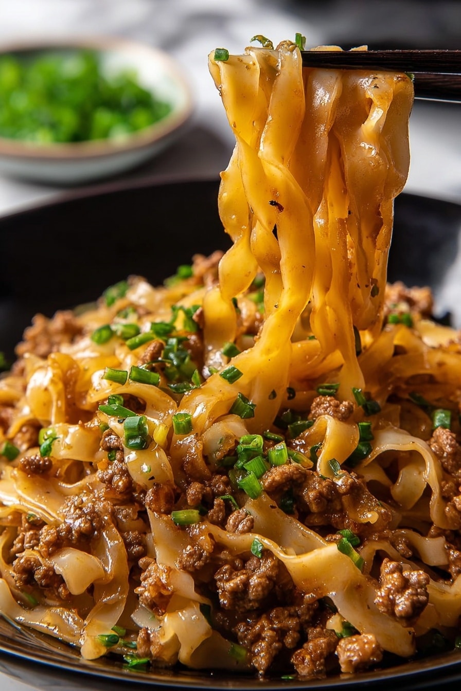 Potsticker Noodle Bowl with Pork and Greens Recipe 6 The image shows a close-up of a black bowl filled with thick, wide noodles mixed with small pieces of cooked ground meat, all coated in a shiny brown sauce. Small bright green chopped chives are sprinkled evenly throughout the noodles, adding color contrast. A pair of black chopsticks lifts a bunch of the noodles from the bowl, showing their soft, smooth texture and the meat clinging to them. In the blurred background, there is a small white bowl with more green chives. The surface below is a white marbled texture. photo taken with an iphone --ar 2:3 --v 7 - Potsticker Noodle Bowl with Pork and Greens, flavorful Asian noodle bowl, quick Asian pork recipe, healthy pork and greens stir-fry, easy weeknight noodle bowl
