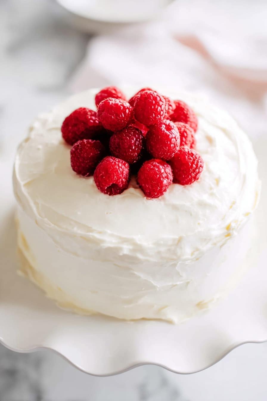 Lemon Raspberry Cake with Cream Cheese Frosting Recipe 6 A round cake with two thick layers covered fully in smooth, white cream frosting is placed on a white plate with wavy edges. On top of the cake is a small pile of fresh, bright red raspberries clustered in the center, adding a pop of color against the white frosting. The cake sits on a white marbled surface with soft, natural light brightening the scene. photo taken with an iphone --ar 2:3 --v 7 - Lemon Raspberry Cake with Cream Cheese Frosting, lemon raspberry cake recipe, moist raspberry cake, creamy frosting cake, citrus berry layer cake