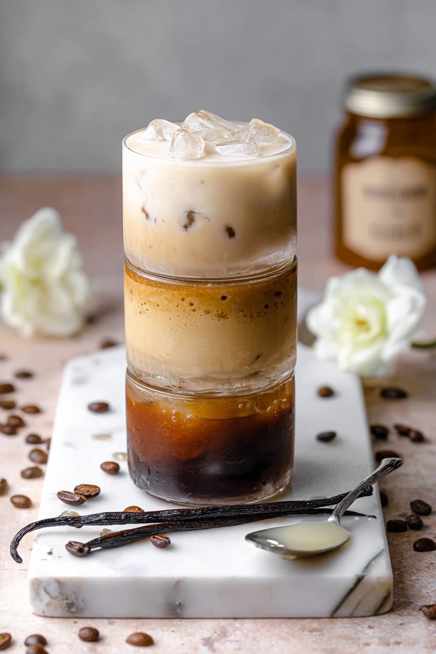 Homemade Vanilla Iced Coffee Recipe 6 The image shows a stack of four iced coffee drinks arranged vertically on a white marbled board. The bottom layer is a dark brown coffee with a smooth, shiny surface. Above it, the second layer is a lighter brown coffee with a subtle ice cube visible inside. The third layer is a creamy beige coffee with small bits floating inside, and the top layer is an almost white cream coffee filled with large ice cubes. Around the stack, there are coffee beans scattered on the white marbled board along with two white flowers and a dark brown vanilla bean pod placed diagonally near the front. A silver spoon with some creamy liquid rests on the side. The background shows soft, neutral tones with a jar blurred in the distance. photo taken with an iphone --ar 2:3 --v 7 - Homemade Vanilla Iced Coffee, vanilla iced coffee recipe, DIY vanilla iced coffee, easy vanilla iced coffee, flavored iced coffee