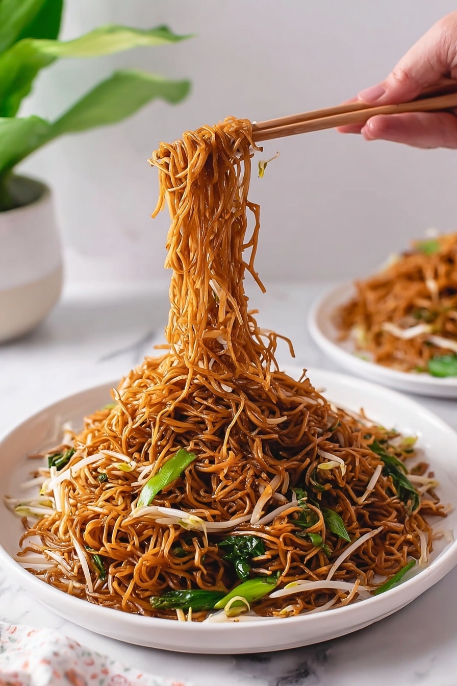 Cantonese Chow Mein Stir Fry Recipe 8 The image shows a white deep plate filled with a large serving of brown stir-fried noodles mixed with white bean sprouts and green leafy vegetables. The noodles are thin and shiny, cooked evenly with a slight oily texture. A woman's hand is using chopsticks to lift a tangled bundle of the noodles from the center, showing the pile of noodles below still resting on the plate. In the background, there is another white plate with more noodles, and a green plant sits next to it, all placed on a white marbled surface. The scene is bright and clean with soft natural light. photo taken with an iphone --ar 2:3 --v 7 - Cantonese Chow Mein Stir Fry, Cantonese Noodle Stir Fry, Chinese Chow Mein Recipe, Crispy Noodles Stir Fry, Vegetarian Chinese Noodles