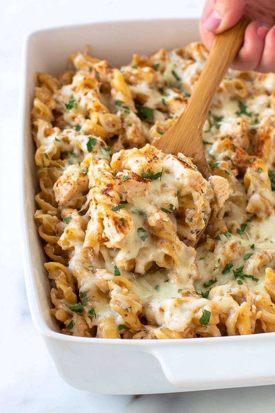Cajun Chicken Pasta Bake Recipe 6 A white rectangular dish filled with a creamy baked pasta dish. The pasta is a mix of short, curly pieces coated in a light brown sauce with melted white cheese spread unevenly on top. Small pieces of cooked chicken are mixed in with the pasta, and a few green herbs are sprinkled across for garnish. A wooden spoon is scooping some of the pasta from the dish, held by a woman's hand. The background is a white marbled surface. photo taken with an iphone --ar 2:3 --v 7 - Cajun Chicken Pasta Bake, Cajun Chicken Pasta, easy Cajun pasta recipes, creamy Cajun pasta, spicy chicken pasta bake