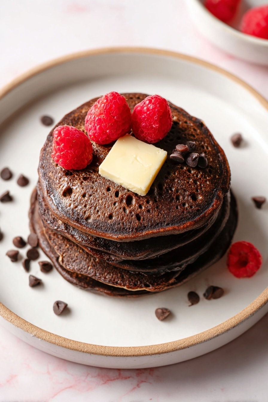 Chocolate Pancakes with Chocolate Chips Recipe 8 A stack of four dark brown chocolate pancakes is placed on a white plate with a thin beige rim, set on a white marbled surface. On the top pancake, there is a square piece of light yellow butter in the center. Three bright red raspberries are arranged around the butter, and small dark brown chocolate chips are scattered on top of the butter and on the plate near the stack. The pancakes have a slightly porous texture with small holes visible. Photo taken with an iphone --ar 2:3 --v 7 - Chocolate Pancakes with Chocolate Chips, easy chocolate pancake recipe, fluffy chocolate pancakes, chocolate breakfast ideas, homemade chocolate pancakes