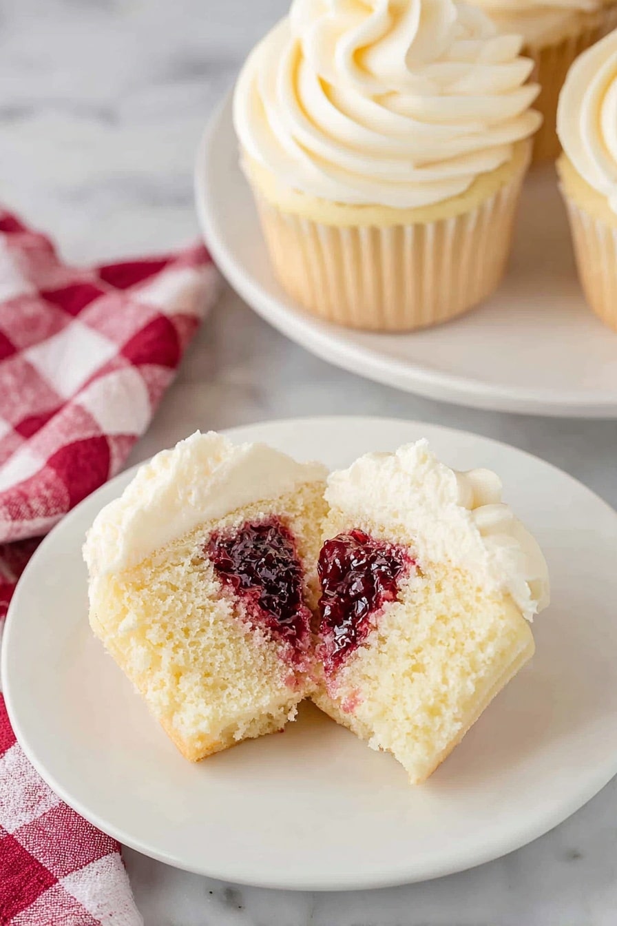 Almond Raspberry Cupcake Recipe 6 A single vanilla cupcake in a white paper liner is shown on a wooden surface, topped with a thick layer of creamy white frosting being piped in a spiral from the outside to the center using a metal star tip. The frosting is smooth and ridged, creating soft swirls with a textured appearance. The background shows a blurred white cloth with red stripes and parts of other cupcakes, with the wooden surface adding warmth to the scene. photo taken with an iphone --ar 2:3 --v 7 - Almond Raspberry Cupcake, almond raspberry cupcakes, raspberry filled cupcakes, elegant cupcake recipes, fruity almond desserts