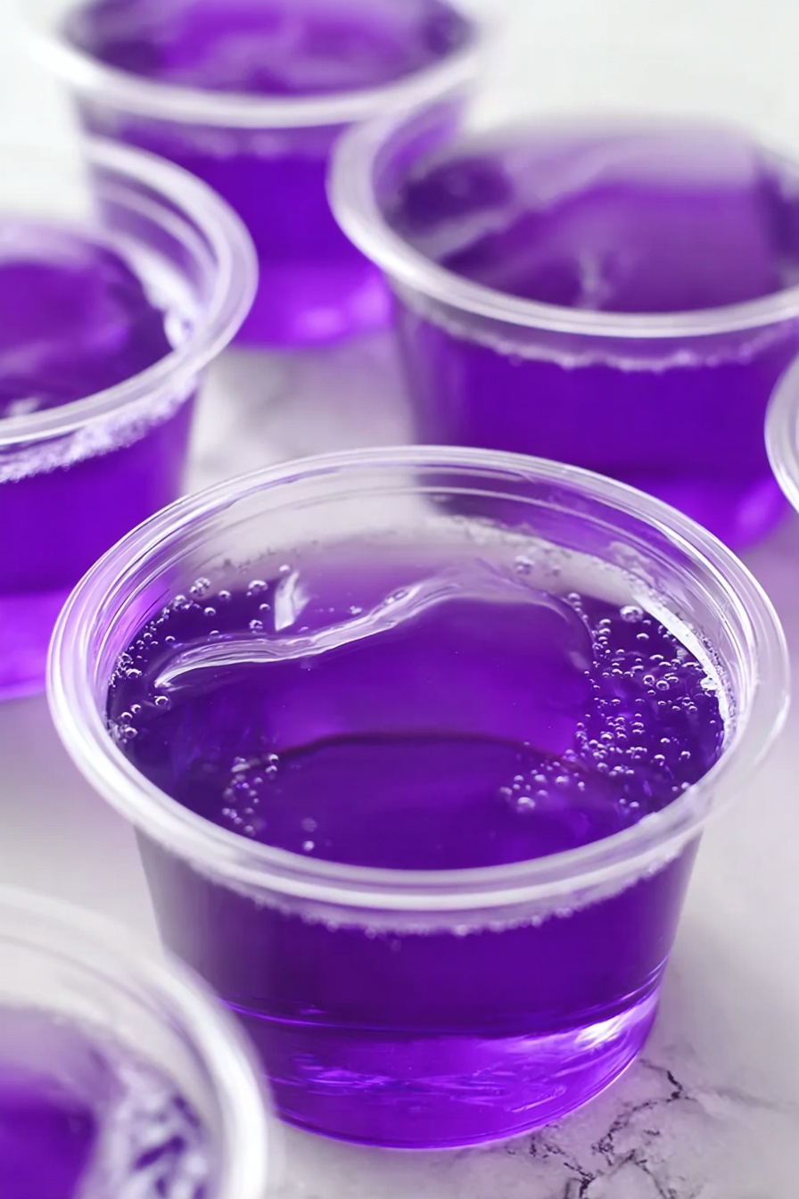 Purple Hooter Jello Shots Recipe 8 The image shows several small clear plastic cups filled with bright purple gelatin. Each cup contains one smooth, shiny layer of gelatin with a few small bubbles and a slightly wavy surface reflecting light. The background is a white marbled texture, softly out of focus, making the vibrant purple color stand out. The cups are close to each other, with one cup in sharp focus in the front center and others blurred in the background. photo taken with an iphone --ar 2:3 --v 7 - Purple Hooter Jello Shots, purple hooter jello shots, layered jello shots with alcohol, party shot recipes, festive alcohol jello shots