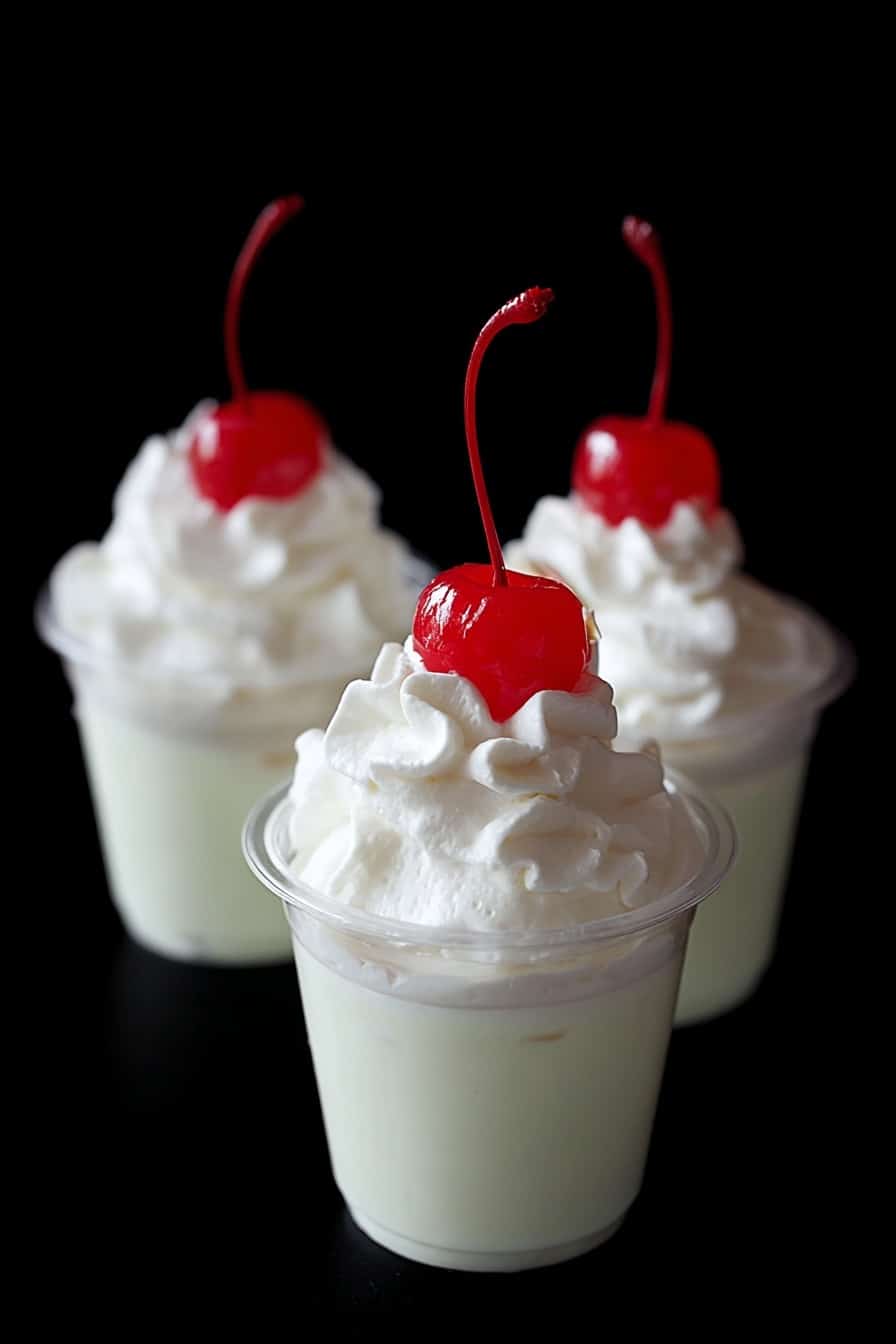 Shamrock Mint Pudding Shots Recipe 6 Three small cups hold a creamy white dessert, each topped with a swirl of white whipped cream and a bright red maraschino cherry with its stem standing up. The cups have two main layers: a smooth, light, pale white base and a fluffy whipped cream layer on top. The background is black, making the white dessert and red cherries stand out clearly. photo taken with an iphone --ar 2:3 --v 7 - Shamrock Mint Pudding Shots, mint pudding shots, St. Patrick's Day dessert, boozy pudding shots, easy party desserts
