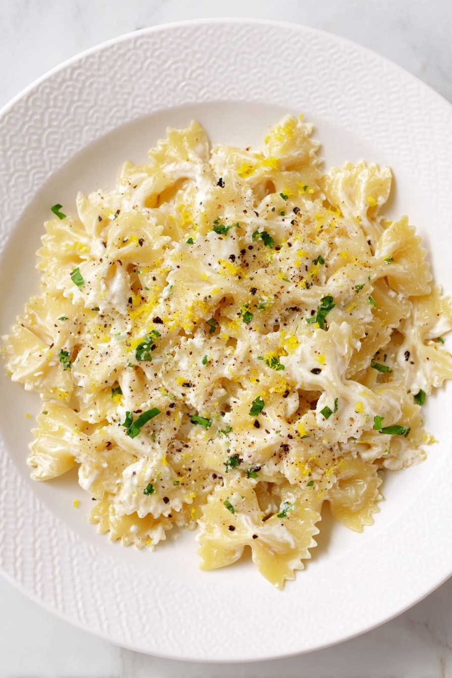 Creamy Lemon Ricotta Pasta Recipe 8 A white plate with a textured edge holds a single layer of farfalle pasta covered with a creamy white sauce that looks smooth and slightly thick. The pasta color is pale yellow, and the sauce is sprinkled with small green herb pieces and black pepper flakes. There is some grated cheese on top, adding a slightly rough texture, and small bits of yellow lemon zest are scattered on the dish, giving a bright contrast. The plate is on a white marbled surface. photo taken with an iphone --ar 2:3 --v 7 - Creamy Lemon Ricotta Pasta, lemon ricotta pasta recipe, quick pasta with ricotta and lemon, easy lemon pasta, bright creamy pasta dish