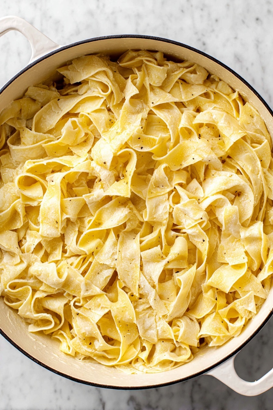 Creamy Lemon Pasta Recipe 8 A large white pot filled with wide, flat pasta strips coated evenly with a creamy, light yellow sauce. The pasta has a soft texture with some pieces slightly curled or folded, showing a mix of smooth and slightly rough edges. The sauce gives a glossy touch, with small flecks of seasoning visible throughout. The pot rests on a white marbled surface, creating a clean and bright background. photo taken with an iphone --ar 2:3 --v 7 - Creamy Lemon Pasta, Lemon Pasta, Easy Lemon Pasta, Bright Pasta Dish, Quick Pasta Recipes