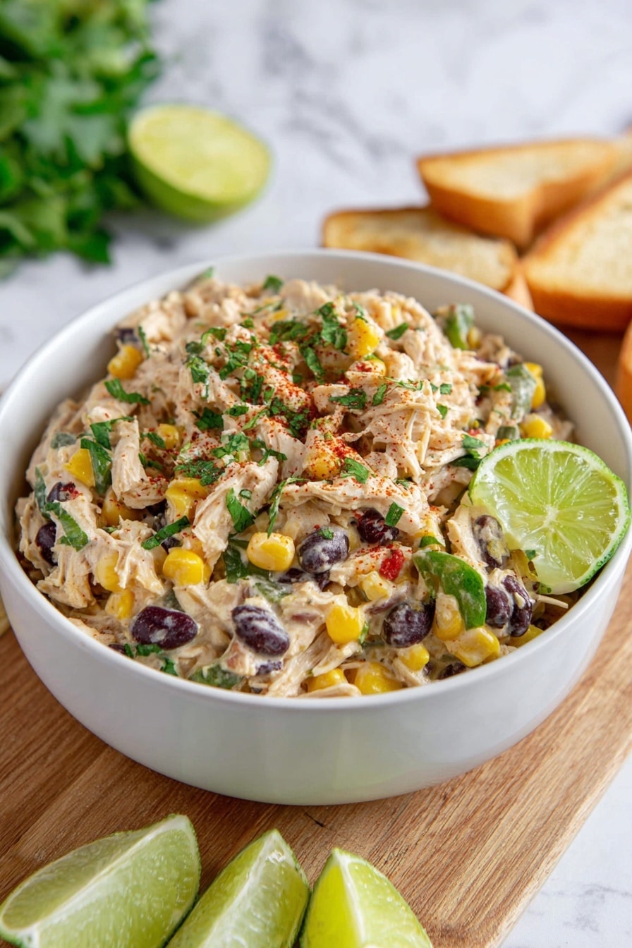 Southwest Chicken Salad Recipe 6 The image shows a white bowl filled with a mixed salad with about three main layers visible: a creamy, light beige base made of shredded light meat, a layer of bright yellow corn and dark black beans scattered evenly, and fresh green bits of chopped herbs and vegetables on top. A wedge of green lime is placed on the edge inside the bowl. The bowl sits on a wooden board with a few lime wedges placed in front and some white toasted bread slices slightly visible behind the bowl. The background has a white marbled texture with blurred green leaves at the top. photo taken with an iphone --ar 2:3 --v 7 - Southwest Chicken Salad, Southwest Chicken Salad Recipe, Easy Southwest Chicken Salad, Healthy Southwest Chicken Salad, Quick Chicken Salad