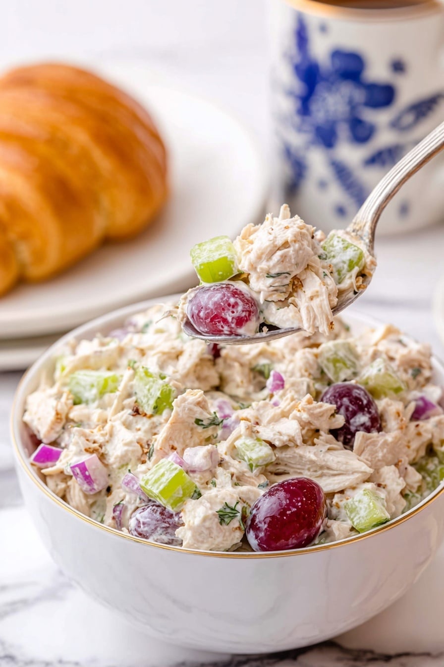 Chicken and Grape Salad Recipe 6 A white bowl filled with a creamy chicken salad showing shredded light-colored chicken mixed with light green celery pieces, small purple-red grape halves, small bright purple onion chunks, and some specks of herbs. A silver spoon is lifting a spoonful of the chicken salad above the bowl, highlighting the colorful mix. In the background, there is a white cup with blue floral designs and a white plate holding a golden croissant, all placed on a white marbled surface. photo taken with an iphone --ar 2:3 --v 7 - Chicken and Grape Salad, Chicken and Grape Salad Recipe, light chicken salad, healthy fruit salad, quick summer salad