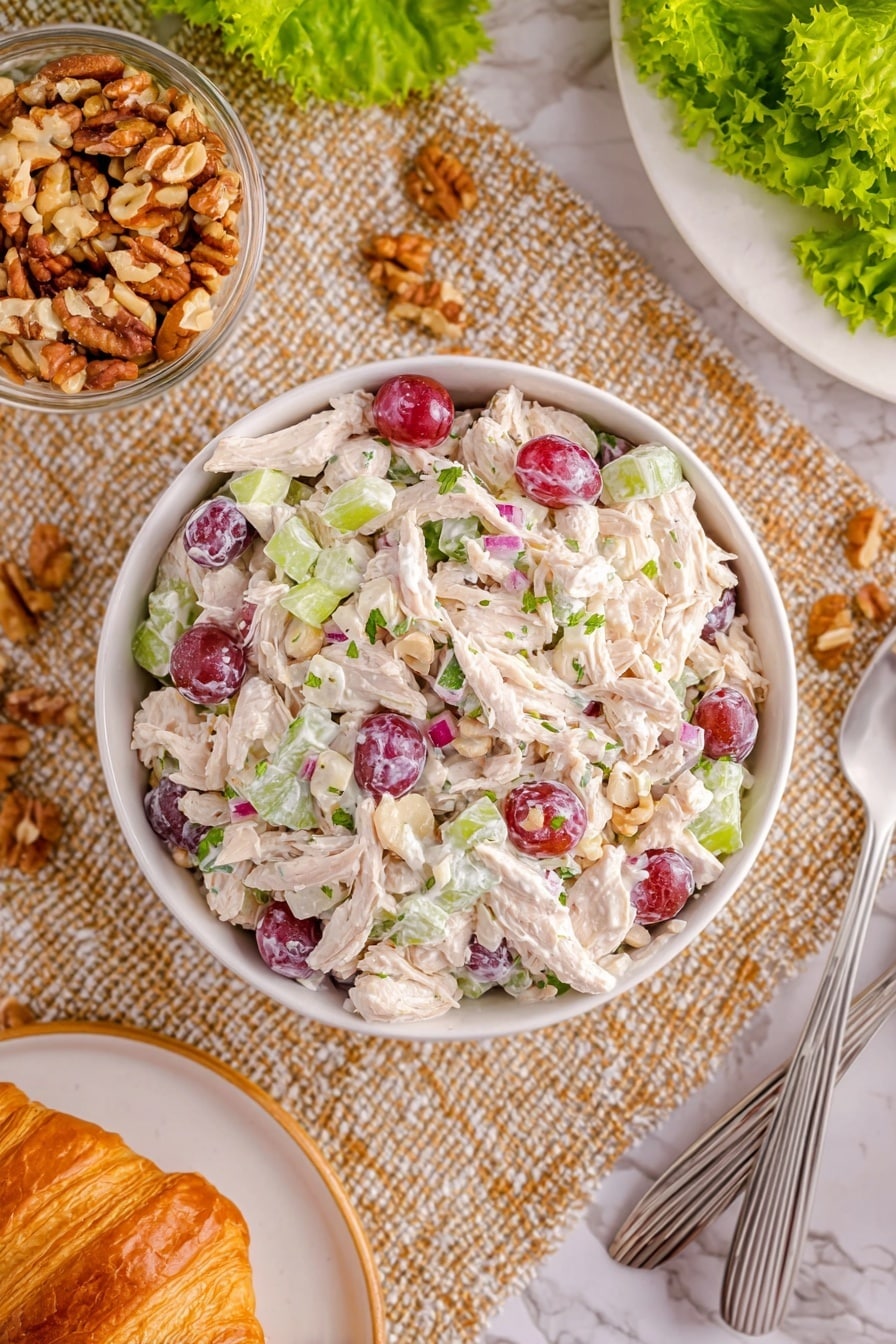 Chicken and Grape Salad Recipe 8 A white bowl filled with a creamy chicken salad layered with thick white shredded chicken pieces, scattered purple-red grape halves, small light green celery slices, and tiny bits of red onion, all mixed with a white creamy dressing with specks of green herbs. The bowl sits on a textured beige woven cloth, surrounded by a glass bowl of chopped light brown nuts, a golden brown croissant on a white plate, two silver spoons, and some bright green lettuce leaves on a white marbled surface. Photo taken with an iphone --ar 2:3 --v 7 - Chicken and Grape Salad, Chicken and Grape Salad Recipe, light chicken salad, healthy fruit salad, quick summer salad