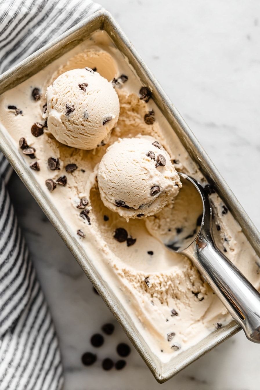 Creamy Cottage Cheese Ice Cream Recipe 6 The image shows a rectangular metal loaf pan filled with light tan ice cream mixed with dark chocolate chips. On top of the ice cream, there are two perfectly round scoops, one closer to the front left and the other near the middle right, both containing visible chocolate chips. A metallic ice cream scooper holding some ice cream rests on the right side of the pan. The pan is placed on a white marbled surface, and a striped cloth is faintly visible in the top left corner. The overall scene looks soft and creamy with a cool, fresh feeling. Photo taken with an iphone --ar 2:3 --v 7 - Creamy Cottage Cheese Ice Cream, healthy cottage cheese ice cream, protein-packed frozen dessert, quick homemade ice cream, low-calorie cottage cheese treat