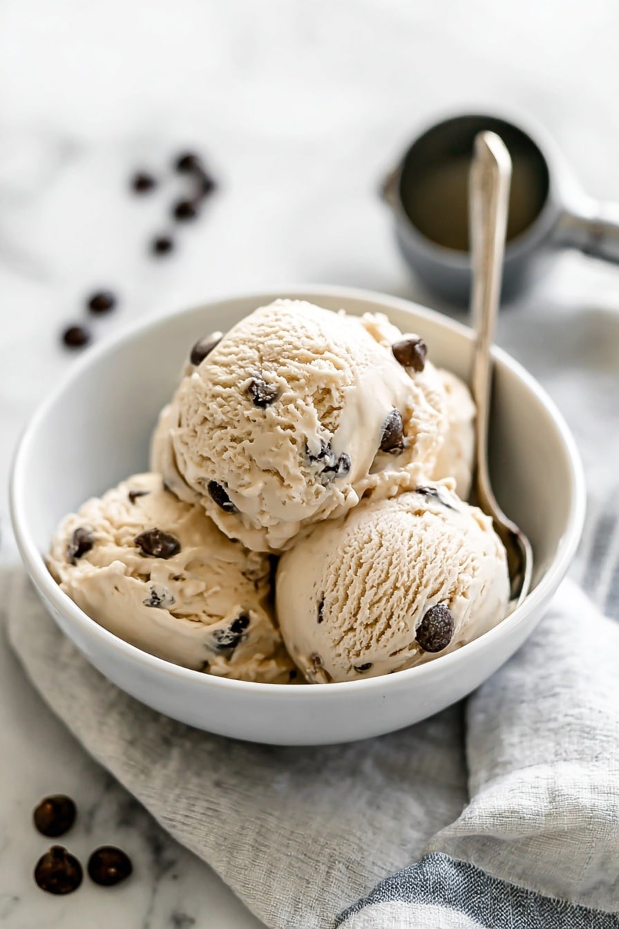 Creamy Cottage Cheese Ice Cream Recipe 8 The image shows a white bowl filled with four scoops of light tan ice cream with dark brown chocolate chips mixed in. The ice cream has a slightly creamy texture and is softly rounded on each scoop, stacked neatly inside the bowl. A metallic spoon with a slim handle rests inside the bowl, pointing outward. The bowl sits on a light cloth with thin stripes, placed on a white marbled surface. In the background, slightly out of focus, there is a metal ice cream scoop and scattered dark chocolate chips. Photo taken with an iphone --ar 2:3 --v 7 - Creamy Cottage Cheese Ice Cream, healthy cottage cheese ice cream, protein-packed frozen dessert, quick homemade ice cream, low-calorie cottage cheese treat
