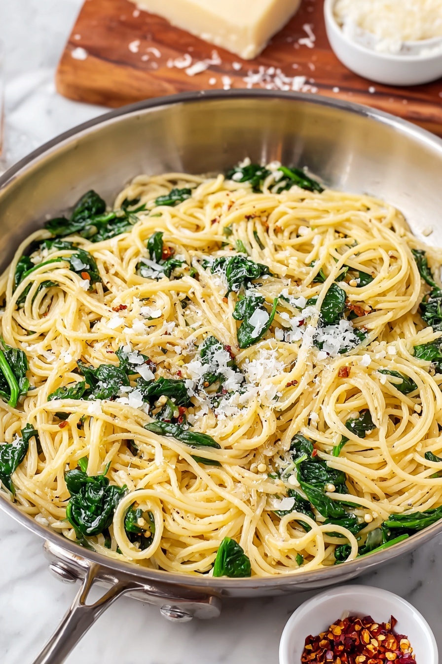 Lemon Spinach Pasta Recipe 6 The image shows a shiny silver pan filled with spaghetti pasta mixed with bright green spinach leaves scattered evenly throughout. The pasta appears soft and is lightly coated with oil or sauce, giving it a glossy, smooth texture. Small bits of grated white cheese are sprinkled on top, adding texture and contrast to the yellowish pasta and green spinach. In the background, there is a piece of cheese placed on a wooden board and a small white bowl filled with red chili flakes on a white marbled surface. The overall look of the dish is fresh, light, and inviting. photo taken with an iphone --ar 2:3 --v 7 - Lemon Spinach Pasta, quick lemon pasta, healthy spinach pasta, citrus pasta recipe, easy vegetarian pasta