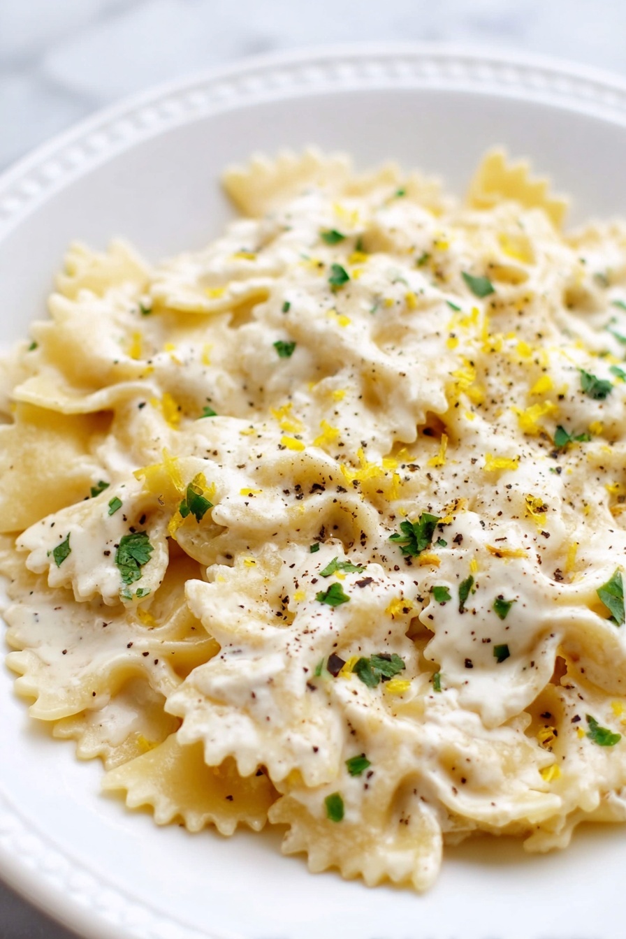 Creamy Lemon Ricotta Pasta Recipe 6 This image shows a white plate with one layer of bow-tie pasta covered in a creamy white sauce with a smooth texture. Sprinkled on top are small bits of green herbs, tiny black pepper pieces, and light yellow zest, adding contrast and color. The sauce covers most of the pasta, making the dish look rich and creamy. The background is a white marbled surface. photo taken with an iphone --ar 2:3 --v 7 - Creamy Lemon Ricotta Pasta, lemon ricotta pasta recipe, quick pasta with ricotta and lemon, easy lemon pasta, bright creamy pasta dish