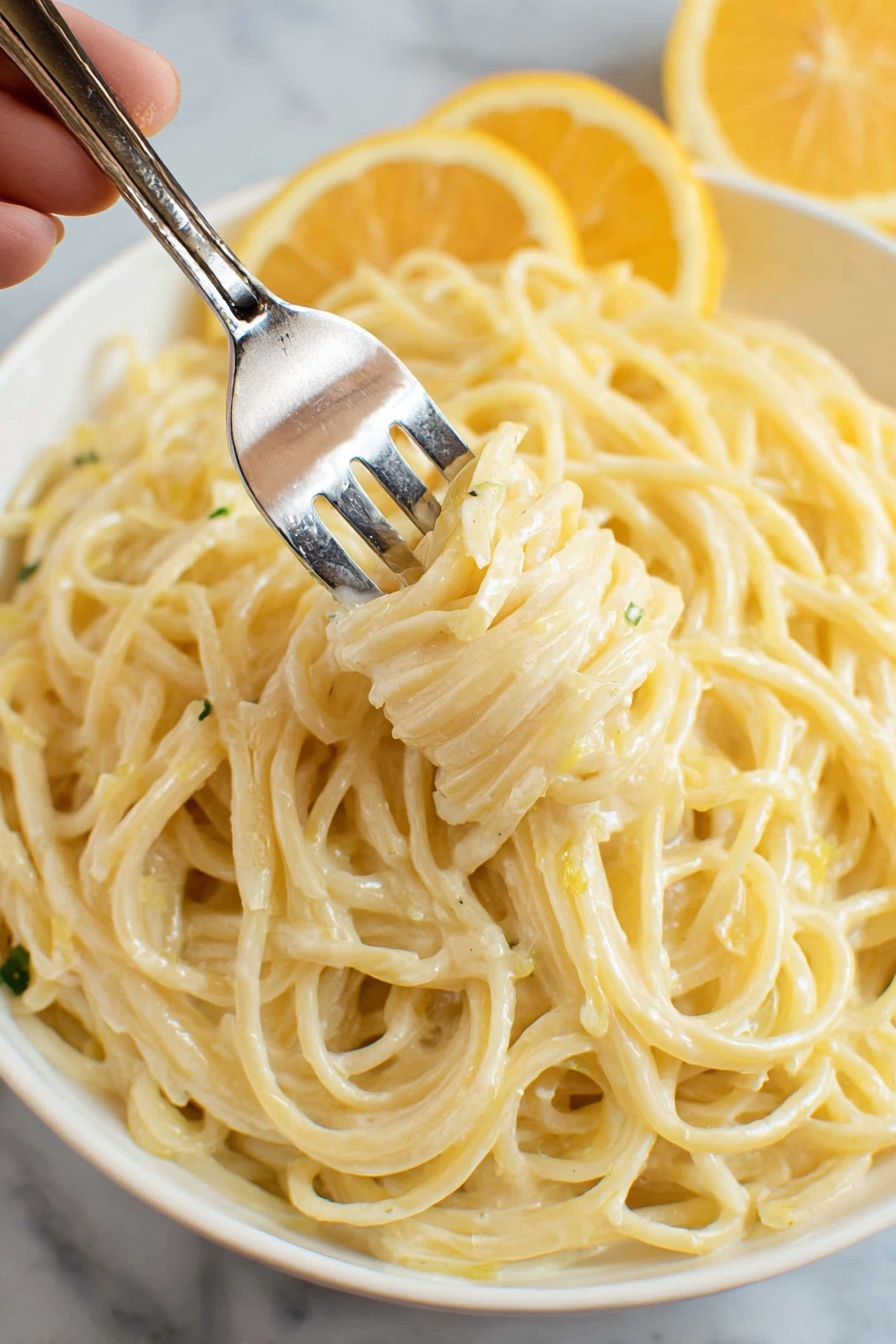 Lemon Garlic Pasta Recipe 8 A close-up image shows creamy spaghetti pasta with a smooth, pale yellow sauce mixed evenly throughout. The noodles are thin and glossy, twisted around a silver fork held by a woman's hand in the upper left. The plate beneath is white, filled with more spaghetti forming an intertwined base layer. In the background on the plate's edge, two lemon wedges add a bright, fresh yellow contrast. The entire scene is set against a white marbled surface. photo taken with an iphone --ar 2:3 --v 7 - Lemon Garlic Pasta, Lemon Garlic Pasta Recipe, easy lemon garlic pasta, quick pasta dinner, flavorful lemon garlic pasta