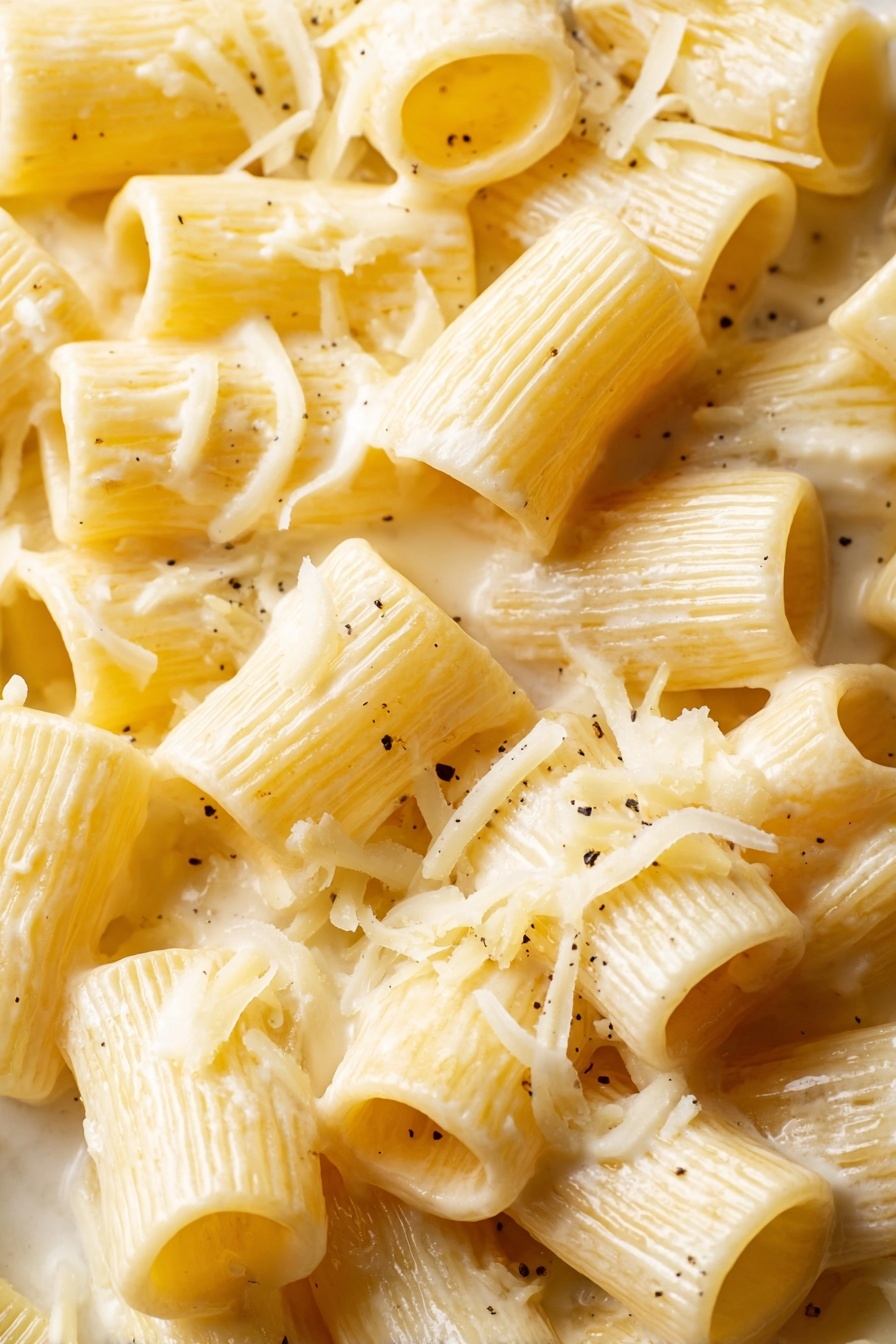 Creamy Lemon Pasta Recipe 6 This close-up image shows many tube-shaped pasta pieces, each light yellow with a smooth, slightly ribbed texture. The pasta is coated in a creamy white sauce that clings to the surfaces and fills some tubes, giving a soft, glossy look. Thin shreds of pale yellow cheese are scattered over the pasta, adding a delicate layered texture. A few tiny black specks are visible, likely black pepper, adding small spots of contrast. The pasta pieces are piled closely together, filling the frame fully, on a white marbled background. photo taken with an iphone --ar 2:3 --v 7 - Creamy Lemon Pasta, Lemon Pasta, Creamy Pasta Recipes, Quick Lemon Pasta, Bright Lemon Pasta