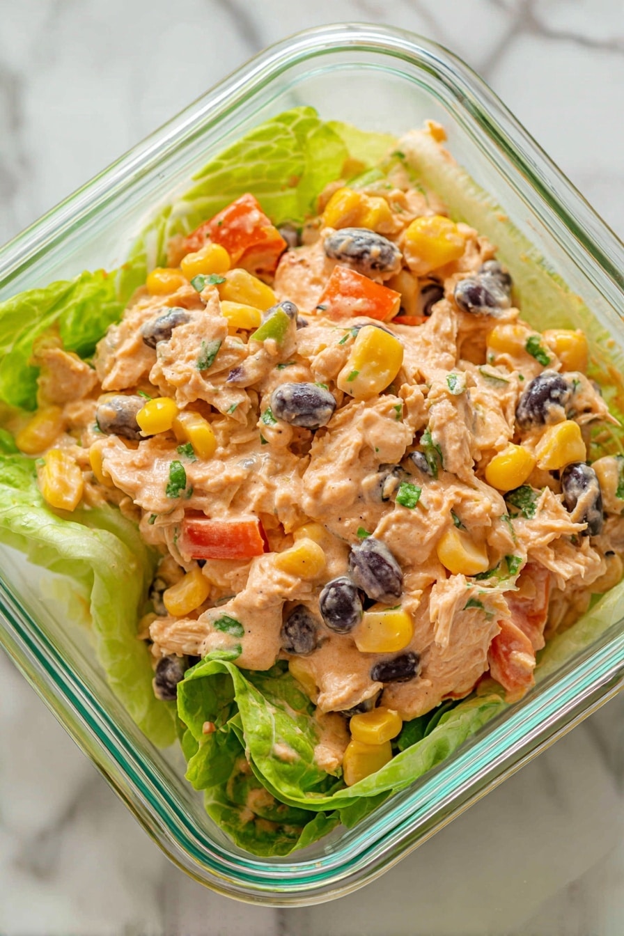 The image shows a dish in a clear glass square container placed on a white marbled surface. The dish has three main layers: a base of light green romaine lettuce leaves, a middle layer of shredded light brown chicken mixed with chunks of bright yellow corn, diced orange bell peppers, and whole black beans, all coated with a creamy, light orange sauce that has a smooth texture. There are small green herb bits scattered throughout the mix. The whole dish looks fresh and colorful with the contrast of the sauce and vegetables against the green lettuce. Photo taken with an iphone --ar 2:3 --v 7 - Chipotle Lime Chicken Salad, spicy chicken salad, healthy chicken salad recipe, quick chicken salad, smoky lime chicken salad