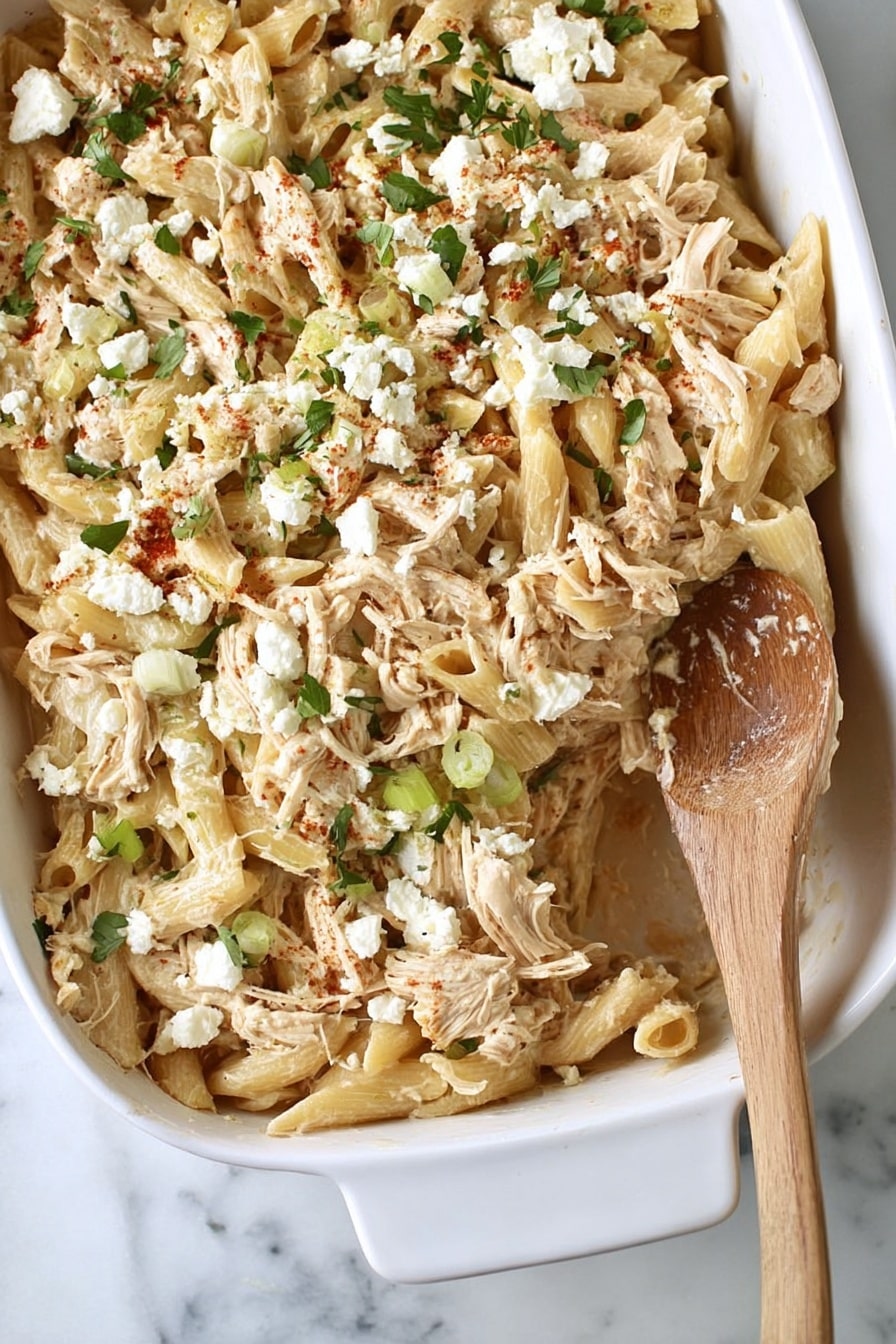 Baked Feta Pasta with Chicken Recipe 6 The image shows a white baking dish filled with a creamy pasta bake. The pasta is light beige and shaped in short tubes mixed with shredded pieces of chicken. Scattered on top are small white clumps of cheese and finely chopped green herbs, adding a fresh contrast. A wooden spoon is resting on the right side of the dish, partly sunk into the pasta. The background is a white marbled surface. photo taken with an iphone --ar 2:3 --v 7 - Baked Feta Pasta with Chicken, easy baked feta pasta, creamy chicken pasta bake, one-pan feta pasta, quick weeknight dinner